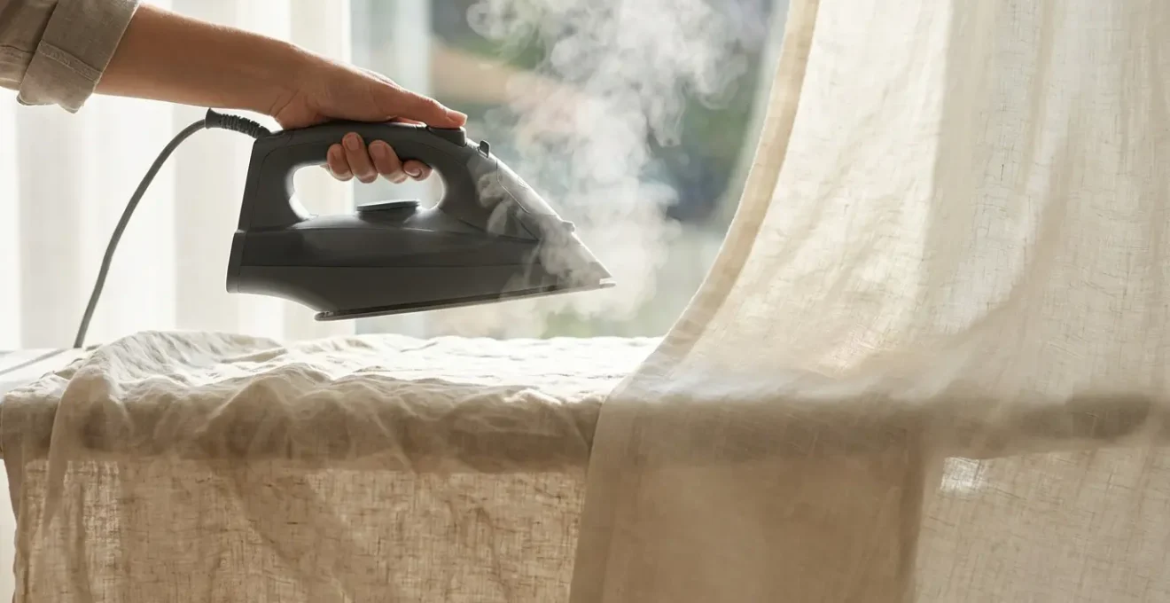 Steam iron hovering over damp linen curtain fabric creating soft steam clouds